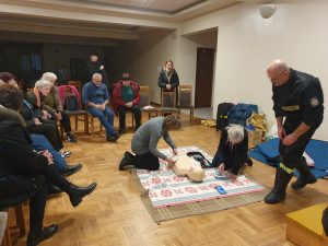 Grupa starszych osób siedzi na sali. Na środku sali dwie kobiety trenują na manekinie udzielanie pierwszej pomocy.