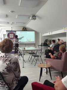 Grupa seniorów siedzi na sali, w oddali wyświetlana jest prezentacja multimedialna