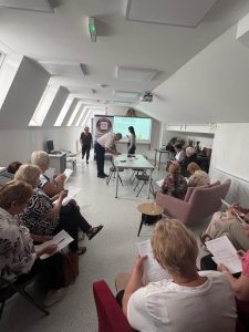 Grupa seniorów siedzi na sali, w oddali wyświetlana jest prezentacja multimedialna