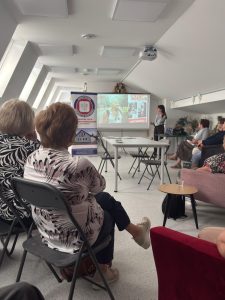 Grupa seniorów siedzi na sali, w oddali wyświetlana jest prezentacja multimedialna