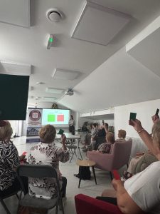Grupa seniorów siedzi na sali, w oddali wyświetlana jest prezentacja multimedialna